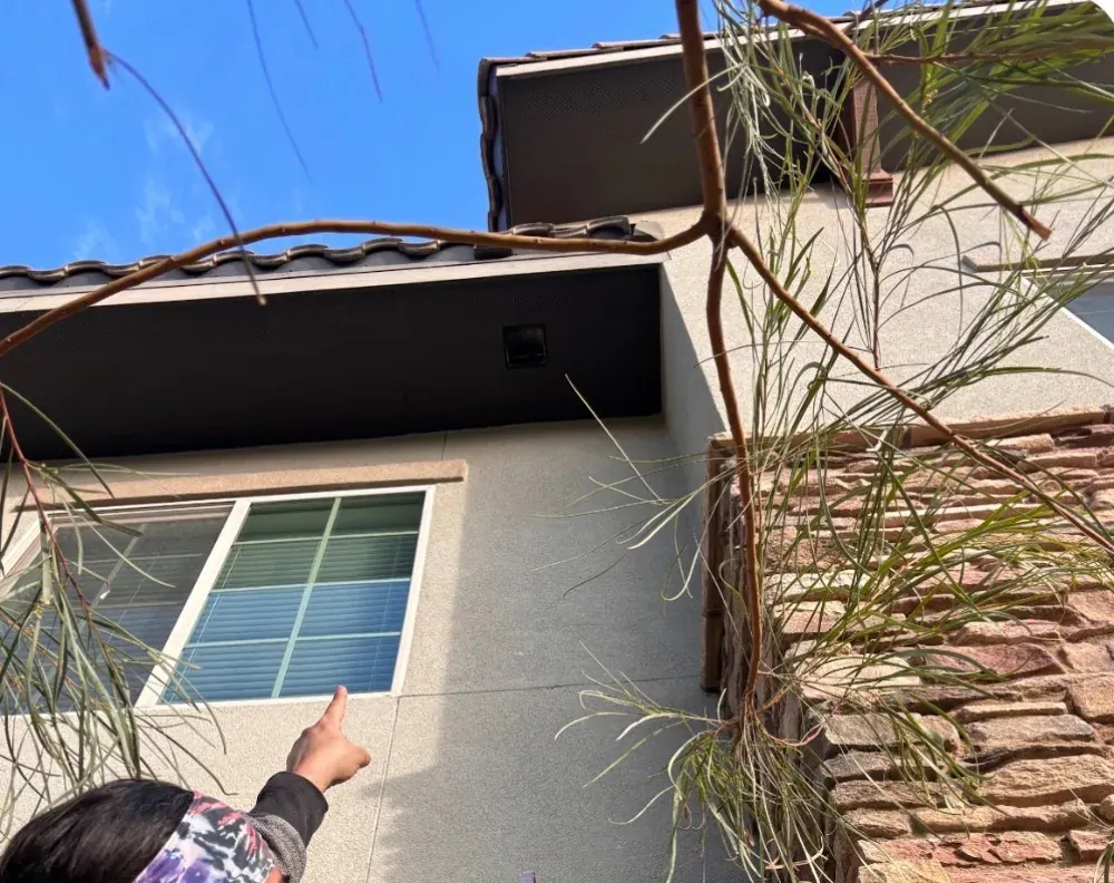Homeowner pointing up at a dryer vent outlet high on a two-story stucco wall under the soffit